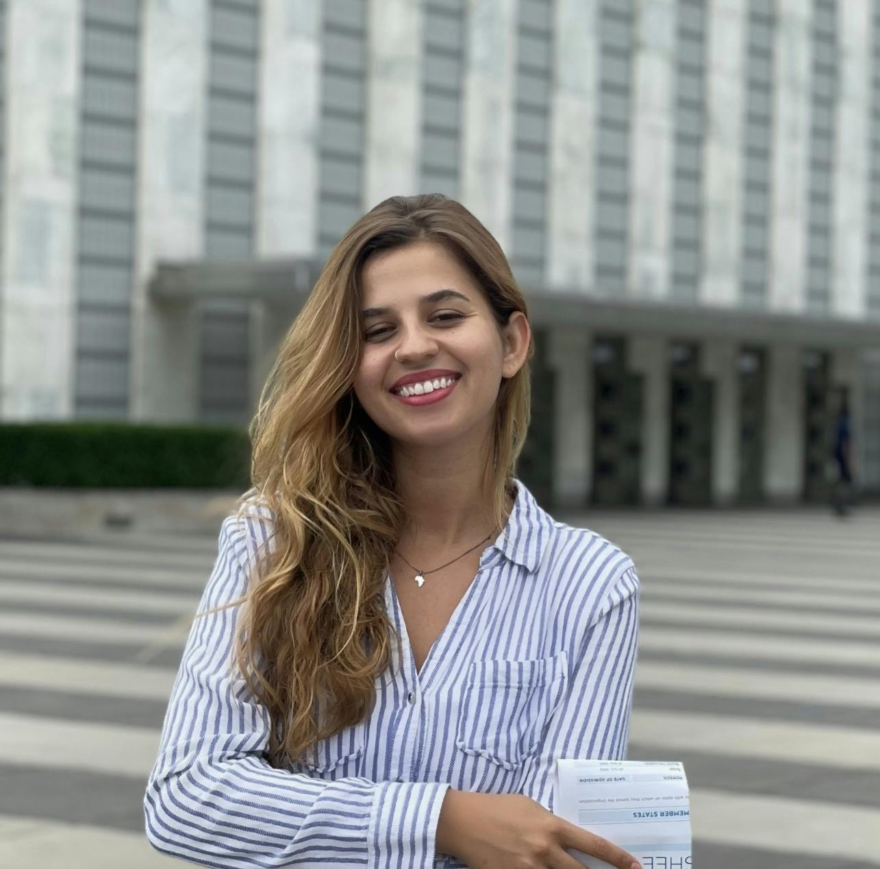 Flávia smiles in front of a large building. She is wearing a blue and white striped shirt.