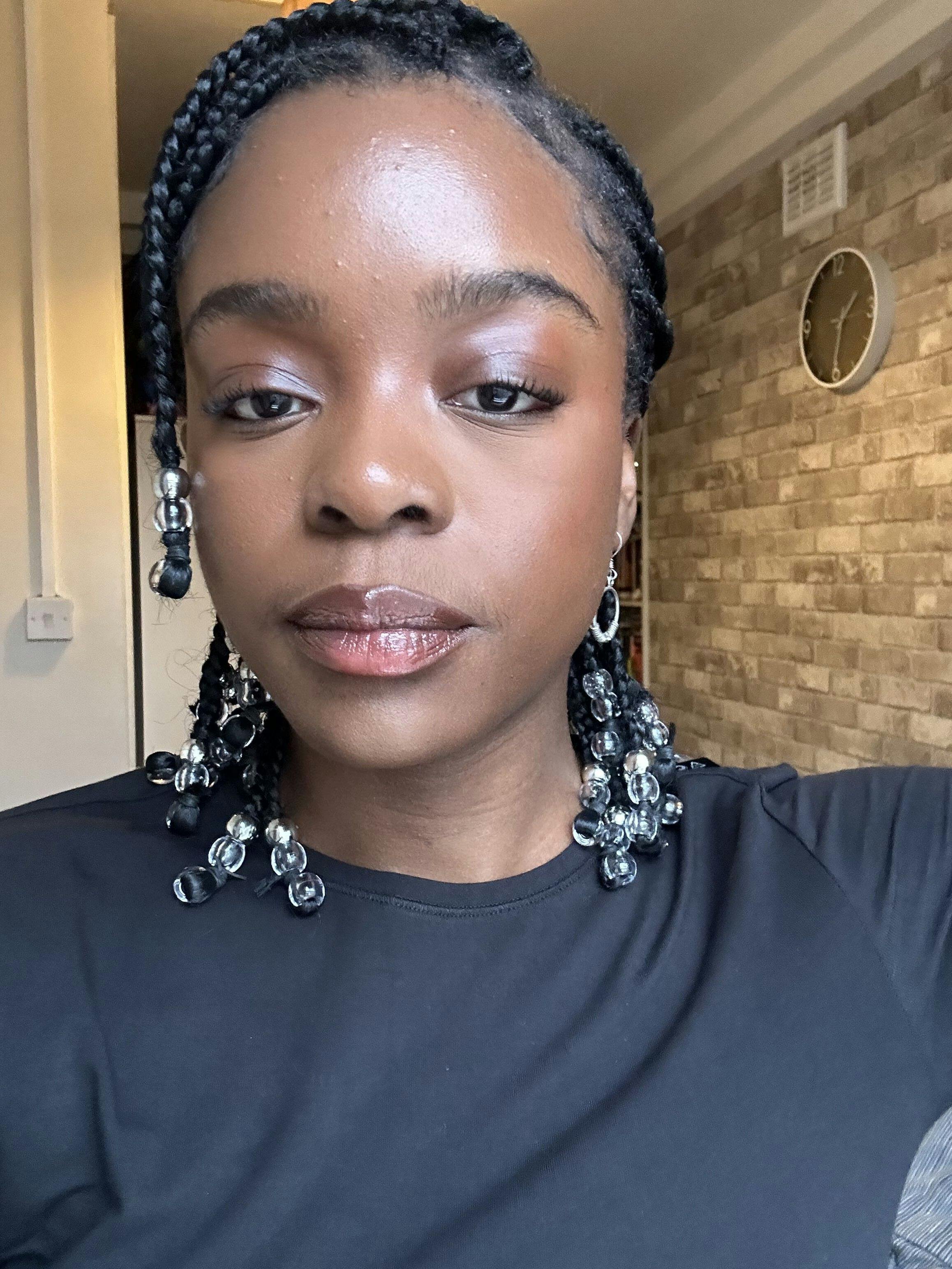 Abbie is a Black woman taking a selfie. She is wearing a black top and has black hair. She is standing in front of a brick wall with a clock on it.